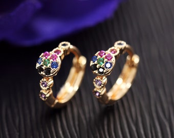 May include: A pair of gold-colored hoop earrings. Each earring features a cluster of colorful gemstones, including red, blue, green, and purple. The earrings have a small, decorative design along the hoop.