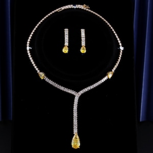Op de afbeelding: Een goudkleurige ketting en oorbellen set. De ketting heeft een delicate ketting met een hanger van een gele druppelvormige steen. De oorbellen passen erbij, met een gele druppelsteen aan de onderkant. De set wordt gepresenteerd op een zwarte ondergrond.