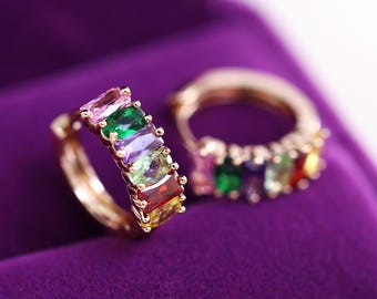 May include: Two gold-colored hoop earrings, each adorned with a row of colorful rectangular gemstones. The gemstones include pink, green, purple, and yellow hues. The earrings are set against a purple background.