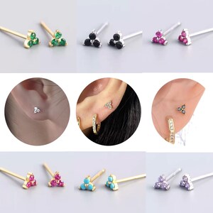 May include: A selection of stud earrings with a triangular arrangement of three gemstones. The earrings come in several colours: green, black, pink, turquoise, and purple. Some earrings are shown worn in ear piercings.