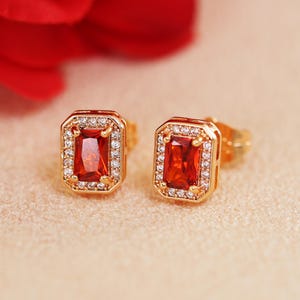 May include: A pair of gold-toned stud earrings. Each earring features a rectangular, red gemstone surrounded by a halo of small, clear crystals. The earrings are set against a soft, neutral background.