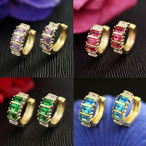 May include: Four pairs of gold hoop earrings with different coloured gemstones. Each pair features three rectangular gemstones with smaller clear stones surrounding them. The colours are purple, red, green, and blue.