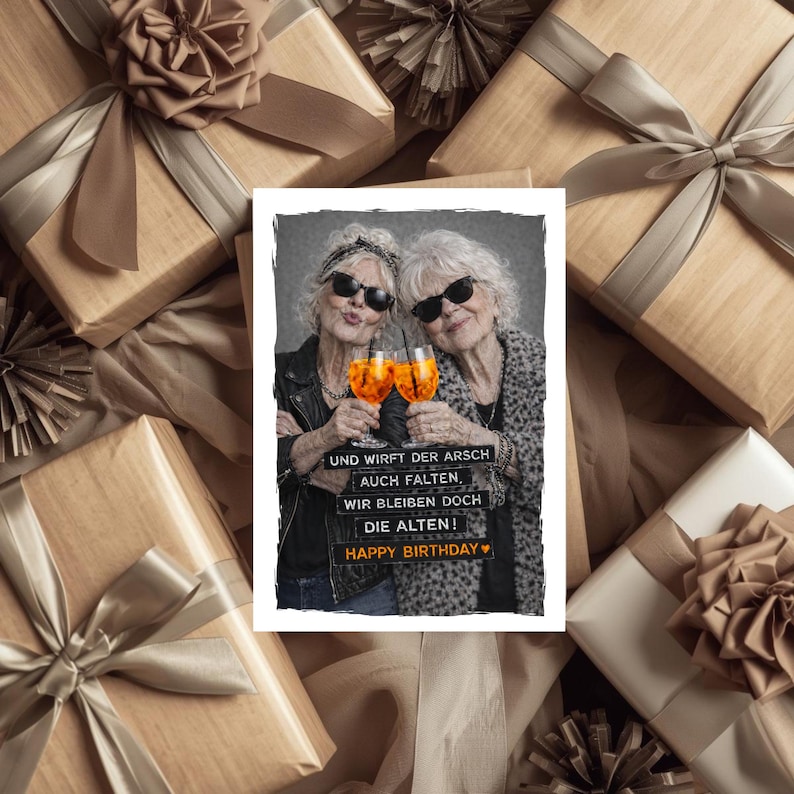 May include: A birthday greeting card featuring two older women wearing sunglasses, holding orange cocktails, and toasting. The card has the text "UND WIRFT DER ARSCH AUCH FALTEN, WIR BLEIBEN DOCH DIE ALTEN! HAPPY BIRTHDAY". The card is surrounded by wrapped gifts.