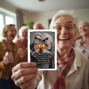 May include: A greeting card with a black and white image of two older women wearing sunglasses, holding orange cocktails, and the text "Happy Birthday". The card is held by a person in front of a group of people.