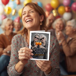 May include: A woman smiles while holding a birthday card with a black and white photo of two older women toasting with orange drinks. The card reads "Happy Birthday" and includes German text.