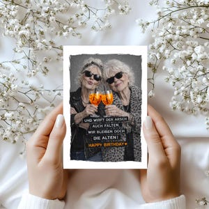 May include: A greeting card featuring two older women wearing sunglasses, toasting with orange cocktails. The card has the text "UND WIRFT DER ARSCH AUCH FALTEN, WIR BLEIBEN DOCH DIE ALTEN! HAPPY BIRTHDAY". The card is held against a background of white flowers.