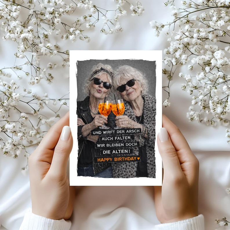 May include: A greeting card featuring two older women wearing sunglasses, toasting with orange cocktails. The card has the text "UND WIRFT DER ARSCH AUCH FALTEN, WIR BLEIBEN DOCH DIE ALTEN! HAPPY BIRTHDAY". The card is held against a background of white flowers.