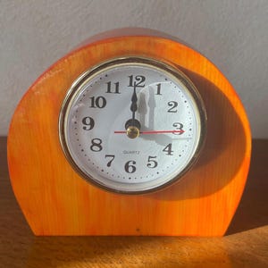 May include: A small, round, analog clock with black hands and a white face. The clock is set to 12:00 and has black numbers. The clock is mounted on a wooden base that is orange and yellow.