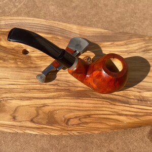 May include: A wooden tobacco pipe with a dark, curved stem and a reddish-brown bowl. A silver and red pipe tool rests beside the pipe. The pipe is set on a wooden surface with a natural grain pattern.