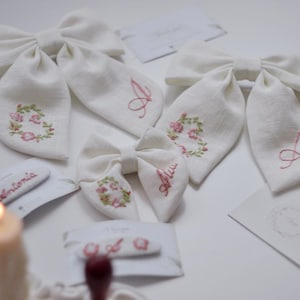 May include: Three off-white fabric hair bows featuring embroidered pink floral wreaths and the cursive letter A in pink thread. Two hair clips are also present, each displaying the name Antonia and the letter A in pink embroidery.
