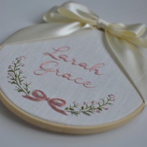 May include: Embroidered hoop art with the name "Larah Grace" in pink script. A floral wreath and bow accent the design on white fabric. A cream-colored ribbon is tied at the top of the wooden hoop.