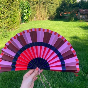 Op de afbeelding: Een handwaaier met een geometrisch patroon van afwisselende segmenten in tinten roze, rood, donkerpaars en diepblauw. De waaier is volledig uitgevouwen in een halve cirkelvorm en heeft een dun, gedraaid rood en wit koord aan de onderkant.