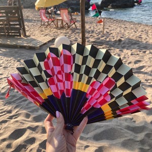 May include: A folding hand fan featuring a black and cream checkered pattern across the top, accented by large pink and red floral shapes. The base of the fan transitions into solid dark blue and yellow sections with a small red cord attached.