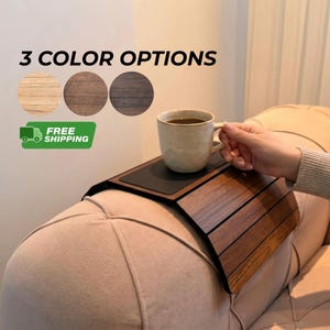 May include: A flexible wooden sofa arm tray with a dark brown finish and a black non-slip center panel. The tray is draped over a beige upholstered sofa arm, supporting a beige ceramic mug. Text reads 3 COLOR OPTIONS.