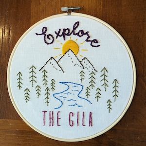 Może przedstawiać: Haft na białym materiale w drewnianym kole z napisem "explore THE GILA". Projekt przedstawia żółte słońce, brązowe góry, zielone drzewa i niebieską rzekę.