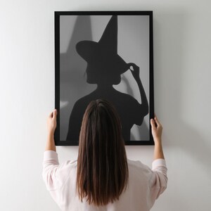May include: A framed black and white print of a witch silhouette. The witch is wearing a tall pointed hat and is touching the brim. The frame is black and the background is a light gray.