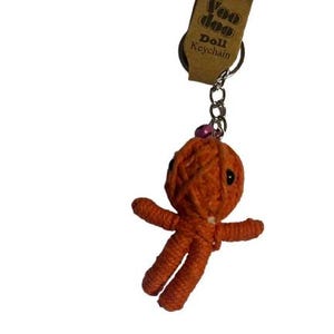 Puede incluir: Llavero de muñeca vudú de hilo naranja con ojos negros y una cuenta rosa. La muñeca tiene los brazos y las piernas extendidos. El llavero incluye una cadena plateada y una etiqueta marrón que dice "Voo doo Doll Keychain".