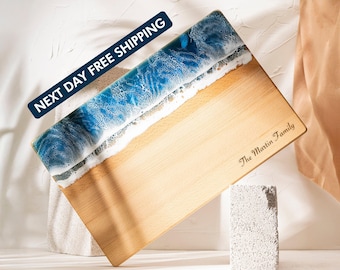 May include: A rectangular wooden cutting board with a beach-themed design. The top edge features a blue and white resin wave design, transitioning to a light brown wooden surface. The text "The Martin Family" is engraved on the board.