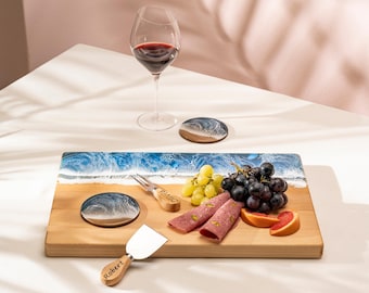 May include: A wooden serving board with a blue and white wave design, paired with a wine glass, coasters, and cheese knives. The board holds grapes, salami, and grapefruit slices. The cheese knives are engraved with names.