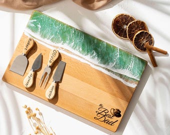May include: Wooden cheese board with a green and white wave design, accompanied by four cheese knives with personalized names. The board is engraved with "the Best Dad" and a hat. Dried orange slices and cinnamon sticks are also present.