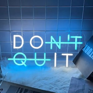 Könnte beinhalten: Ein Neon-Schild mit den Worten "DON'T QUIT" in weißen Neon-Buchstaben mit einem blauen Neon-Umrandung. Das Schild befindet sich auf einer weißen, flauschigen Oberfläche.