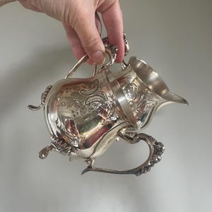 May include: A silver creamer with detailed floral patterns and a curved handle. The creamer features a pouring spout and an ornate handle. The silver surface reflects light, emphasizing the intricate design. The creamer is held by a hand.