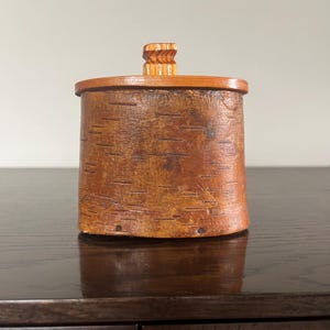 Puede incluir: Una pequeña caja de madera de forma ovalada con un exterior de corteza de abedul. La caja tiene una tapa de madera simple con una parte superior tallada.