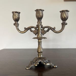 Peut inclure: Un candélabre en laiton à trois branches présentant des détails ornementaux en volutes le long des bras et sur la tige centrale. La base est large et décorative, soutenant trois bougeoirs en forme de tulipe avec des textures nervurées.