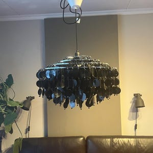 May include: A contemporary chandelier with a layered design, composed of many black, circular, reflective components. The light fitting is suspended from a white ceiling mount with black wiring. The chandelier hangs above a brown leather sofa.