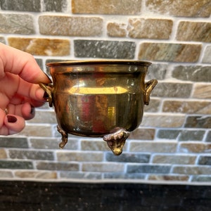 Puede incluir: Un pequeño macetero de metal dorado con dos asas y tres patas. El macetero está vacío y parece ser de latón.