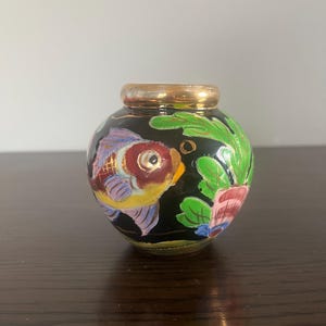 Könnte beinhalten: Kleine, runde schwarze Vase mit goldenem Rand. Die Vase hat ein farbenfrohes, gemaltes Design eines Goldfisches und Wasserpflanzen. Der Fisch hat rote, lila und gelbe Details. Die Pflanzen sind grün und rosa.
