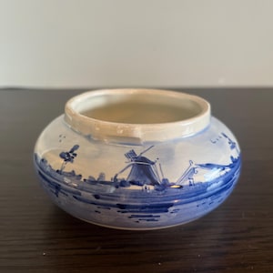 Puede incluir: Un pequeño tarro redondo de cerámica blanca con un diseño pintado en azul y blanco de un molino de viento y una escena de río. El tarro tiene un diámetro de aproximadamente 7,5 cm.