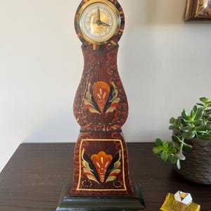 Puede incluir: Un reloj de pie de madera alto con un acabado marrón oscuro y motivos florales pintados a mano en naranja, amarillo y negro. El reloj cuenta con una esfera circular de tono dorado con manecillas y números negros en una estructura curva.