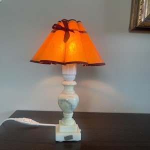May include: A small, illuminated table lamp with an orange lampshade and a marble-like base. The lampshade has a dark red trim and a matching bow. The lamp is turned on, emitting a warm glow. The base is square and has a white cord with an on/off switch.