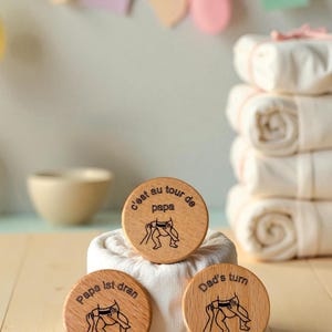 May include: Three round wooden coasters with a baby changing illustration and text. One coaster reads "Dad's turn", another reads "Papa ist dran", and the third reads "C'est au tour de papa". The coasters are light brown and sit on a white surface.