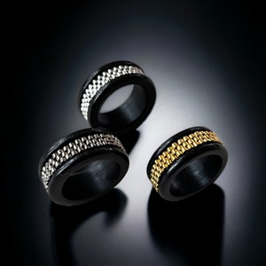 Puede incluir: Tres anillos negros con una banda de metal plateado o dorado. Los anillos se exhiben sobre una superficie oscura y reflectante. Las bandas metálicas tienen una apariencia texturizada, similar a una cadena, que añade un toque de elegancia a la joyería.