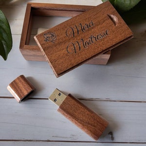 May include: A wooden USB flash drive and matching box. The box lid is engraved with the words "Merci Maîtresse" and a rose. The USB drive is open, revealing the gold-colored connector. The wood has a warm, brown tone.