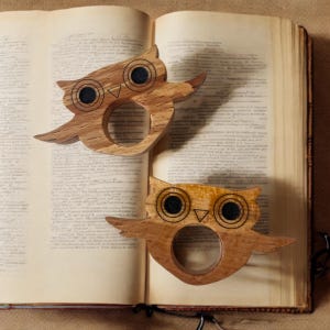 Op de afbeelding: Twee houten uilvormige servetringen. De ringen zijn gemaakt van lichtbruin hout met donkerbruine en zwarte accenten. De uilen hebben grote, ronde ogen en zitten op een open boek. De vleugels zijn uitgespreid en het ontwerp is gedetailleerd.