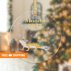 Peut inclure: Une boule de Noël en verre transparent avec un ruban doré et un gecko jaune et noir sur un fond de flocons de neige blancs. La boule indique "Mondo 2023".