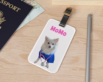 May include: White luggage tag with a black strap and gold buckle. The tag features a white dog wearing a blue shirt and a stethoscope. The name "MoMo" is printed in pink. A silver pen and passport are in the background.