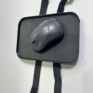 Könnte beinhalten: Ein schwarzes Mauspad mit zwei schwarzen Bändern, die an den Seiten befestigt sind. Das Mauspad ist so konzipiert, dass es an einem Stuhl oder einer anderen Oberfläche befestigt werden kann. Eine schwarze kabellose Maus liegt auf dem Mauspad.