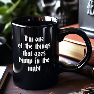 May include: A black ceramic mug featuring white text that reads, I'm one of the things that goes bump in the night. The mug has a standard handle and a cylindrical body, positioned centrally in the frame with a glossy finish on the exterior surface.