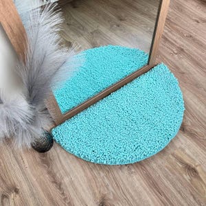 Puede incluir: Una alfombra redonda de color azul claro con una textura esponjosa. La alfombra tiene forma de semicírculo y se coloca delante de un espejo.