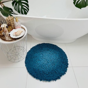 Può includere: Un tappetino da bagno rotondo di colore blu turchese con una superficie testurizzata e soffice. Il tappetino è posizionato su un pavimento piastrellato bianco davanti a una vasca da bagno bianca.