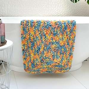 Puede incluir: Una colorida alfombra de baño con un diseño texturizado y en bucle. La alfombra presenta una mezcla de tonos azules, amarillos y naranjas. Está colocada sobre el borde de una bañera blanca, mostrando su apariencia afelpada y su vibrante paleta de colores.