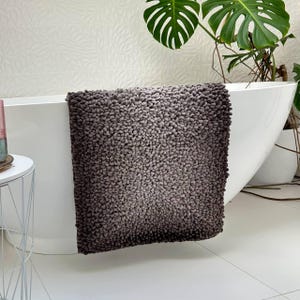 Può includere: Un tappetino da bagno grigio e soffice con una superficie strutturata ad anello è appoggiato sul bordo di una vasca da bagno bianca. Il tappetino ha un effetto sfumato, che passa da un grigio più chiaro a una tonalità più scura. L'ambiente del bagno comprende una parete bianca e una pianta in vaso.