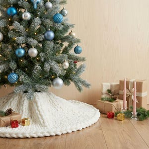 May include: A decorated Christmas tree with blue, silver, and gold ornaments, and a white knitted tree skirt. Presents wrapped in brown paper and candles are arranged on the wooden floor, creating a festive holiday scene.