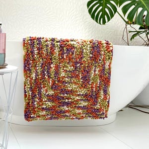 Op de afbeelding: Een kleurrijk badmatje met een getextureerd oppervlak, bestaande uit oranje, paarse, groene en witte plukjes. De vierkante mat is over de rand van een witte badkuip gehangen, geschikt voor een badkamer.