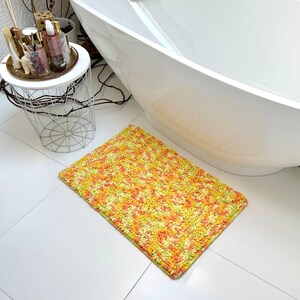 Puede incluir: Una alfombra de baño rectangular con un patrón moteado en tonos de amarillo, naranja y verde. La alfombra está colocada sobre un suelo de baldosas blancas cerca de una bañera blanca. Los bordes de la alfombra son de color verde oscuro.