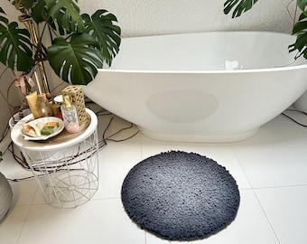 May include: A round, dark gray bath mat with a textured surface sits on white tile flooring. The mat is in front of a white, oval-shaped bathtub. The bathroom has a modern design with a small side table and potted plants.